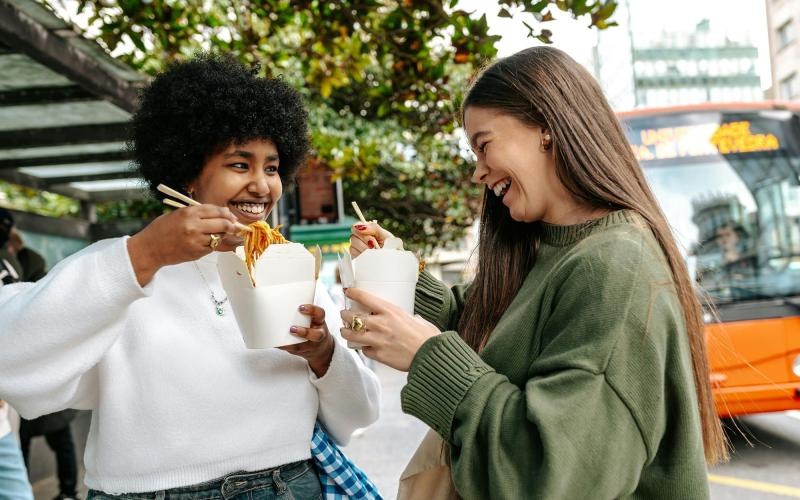 Two women laughing and eating street food in the city.